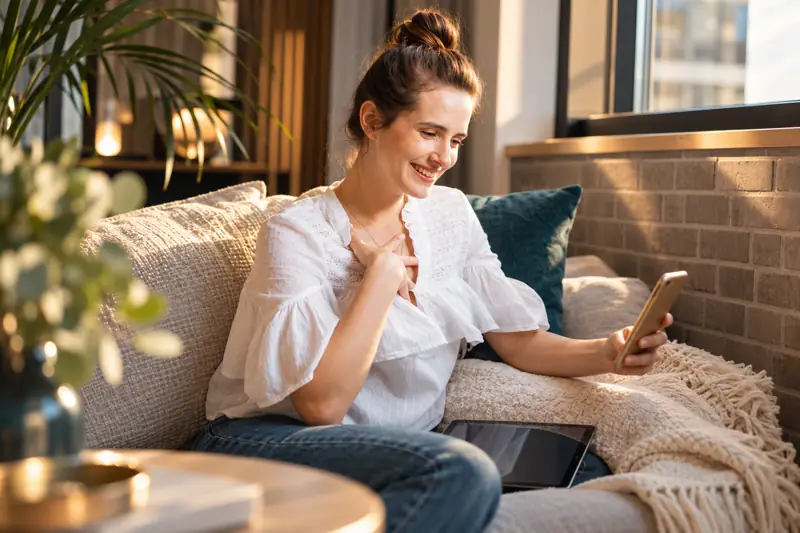 Lächelnde Urlauberin nutzt die Gäste-App auf dem Smartphone auf dem Sofa